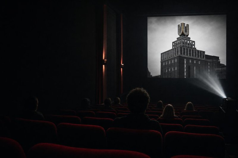 Dunkler Kinosaal mit roten Sitzreihen und Publikum von hinten, auf der Leinwand ein Schwarzweißbild eines Backsteinturms mit einem großen U auf dem Dach (mit AI erstelltes Bild)
